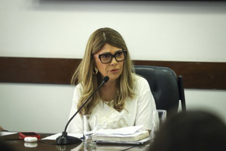 Imagem colorida mostra Cris Monteiro, mulher branca, de cabelos loiros, em uma mesa de reunião, falando ao microfone - Metrópoles