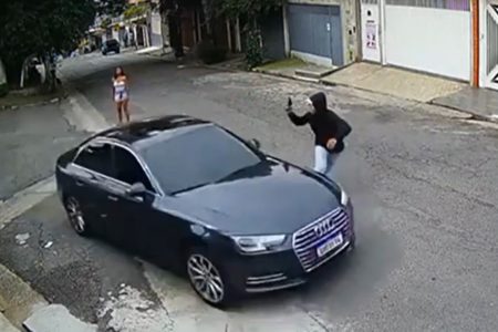 Imagem colorida mostra um homem de blusa preta com capuz apontando uma arma a um carro de luxo de preto; atrás deles, uma mulher vestida de branco e short jeans, aparentemente a isca para o chamado "golpe do amor" - Metrópoles