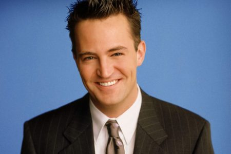 Foto colorida de Matthew Perry vestido de terno com fundo azul - Metrópoles