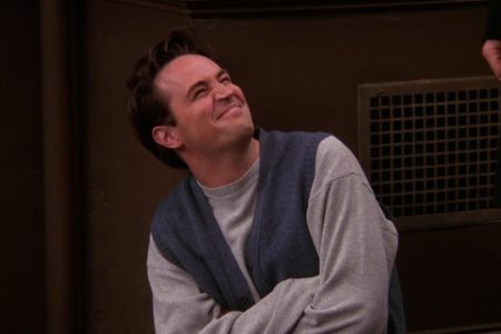 Matthew Perry como Chandler Bing com os braços cruzados vestindo camisa e colete cinzas - Metrópoles