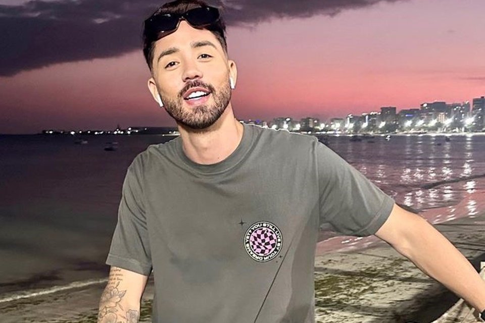 Rico Melquiades posa de óculos escuros na cabeça e blusa cinza na orla - Metrópoles