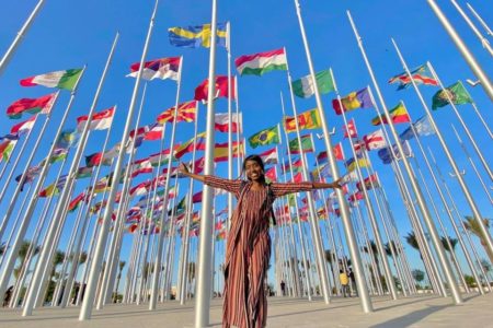 Foto colorida da influencer Nataly Castro com os braços abertos, usando vestido estampado e com bandeiras de diversos países como fundo - Metrópoles