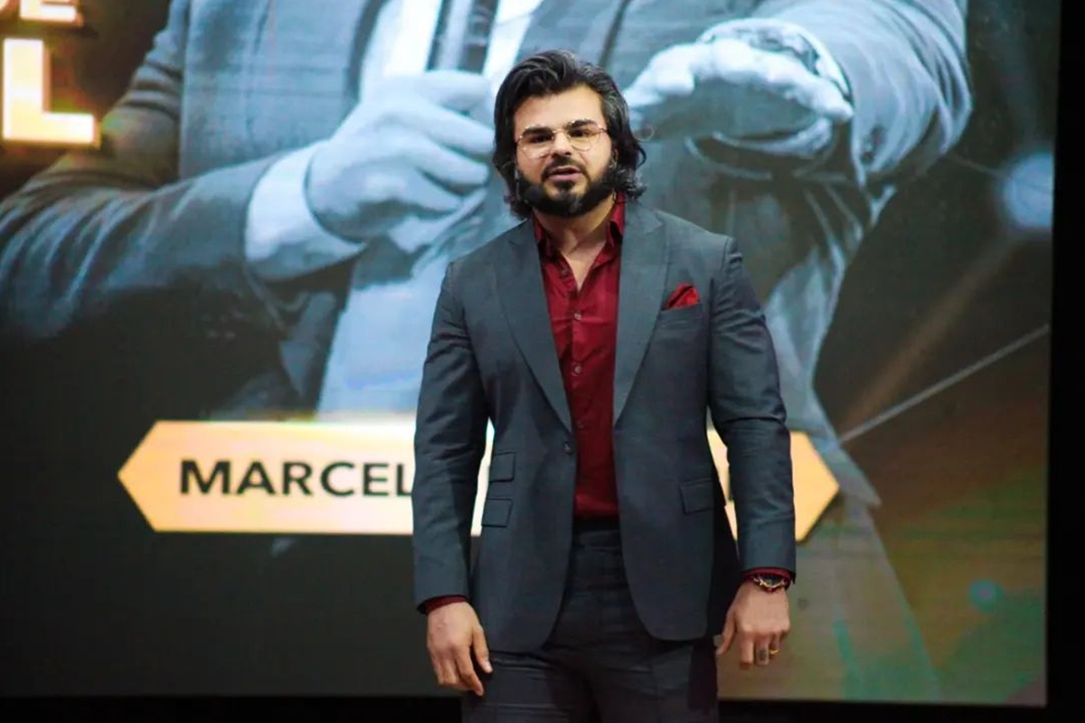 Foto colorida de homem com blazer preto e blusa vermelha em frente um backdrop - Metrópoles