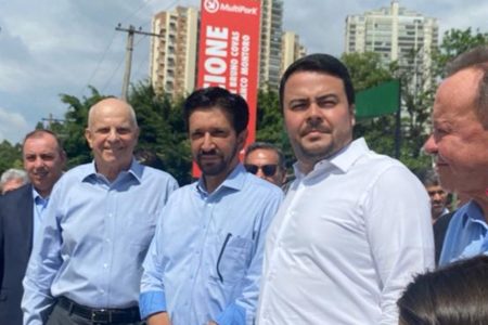 Imagem colorida mostra Ricardo Nunes, chefe de Prefeitura de São Paulo, um homem branco de cabelo e barba pretos, de camisa azul, entre dois homens vestidos de forma parecida, sob sol forte, na rua - Metrópoles