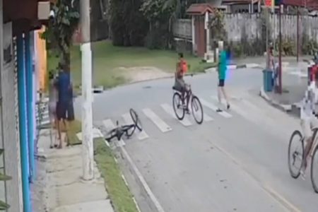 Imagem colorida mostra 3 bandidos de bicicleta abordando idosa no litoral de São Paulo. Um deles encosta a mulher em um muro de uma casa para roubar uma corrente - Metrópoles