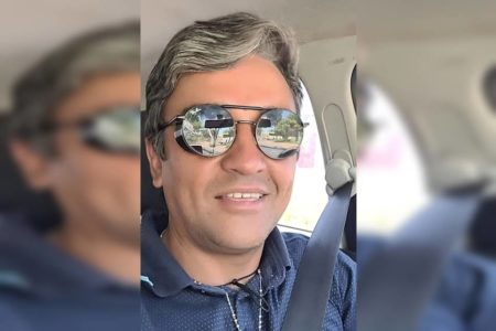 Homem branco com cabelo grisalho e óculos escuros que refletem, blusa azul gola polo