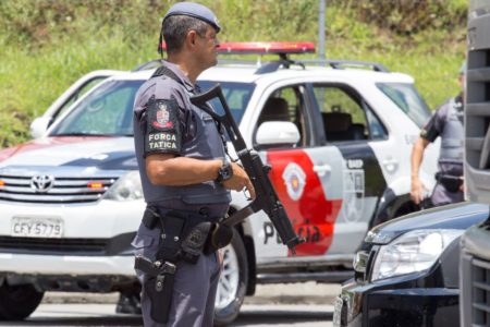 Foto colorida de policial militar armado com viatura da PM ao fundo - Metrópoles