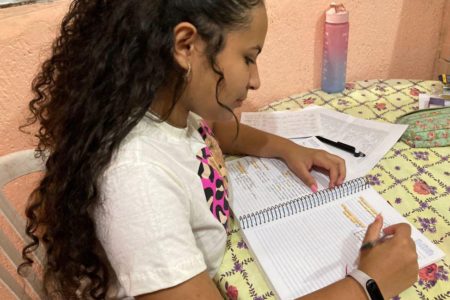 Imagem colorida mostra Deborah Maria da Silva Santos, uma mulher de pele morena, cabelo comprido cacheado preto, usando uma camiseta branca, segurando uma caneta e olhando para um caderno. Ela é uma das estudantes que vai fazer o Enem em 2023 - Metrópoles