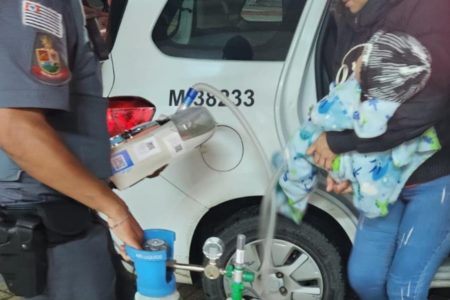 Imagem colorida mostra policial militar paulista segurando um aparelho respiratório, com cilindro de oxigênio, ao lado de um carro da PM. Na frente do policial, há uma mulher, de calça jeans e blusa escura, segurando um bebê. A imagem não mostra o rosto deles - Metrópoles