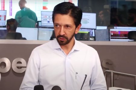 Imagem colorida mostra Ricardo Nunes, homem branco de cabelo e barba pretos, de camisa branca, em um púlpito com microfones. Ele está em uma sala de controle de energia da Enel - Metrópoles