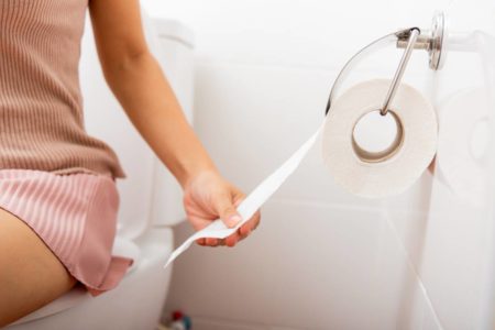 Imagem colorida de mulher sentada no vaso sanitário pegando papel higiência para se limpar - Metrópoles