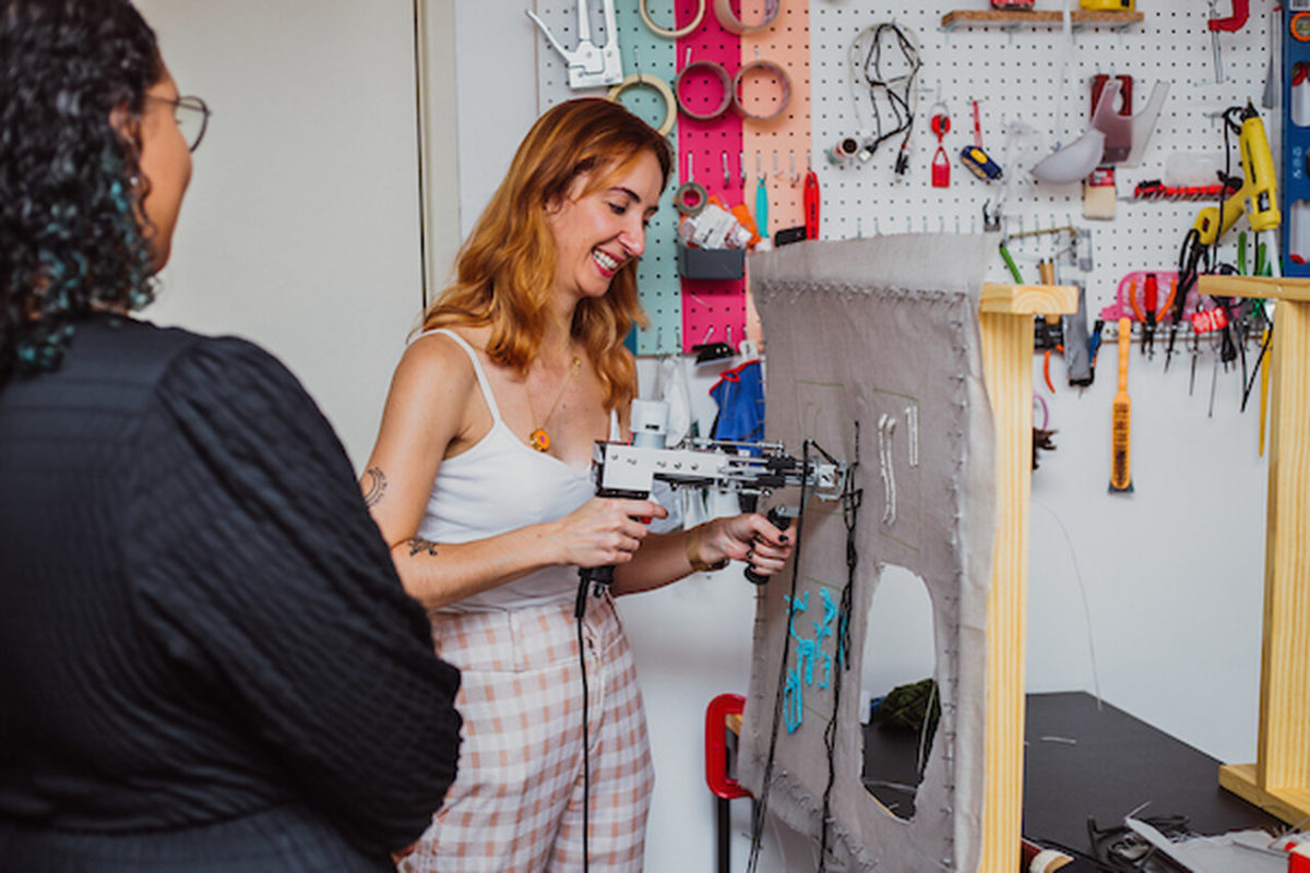 Na imagem com cor, uma mulher branca, ruiva, com roupas claras, usando uma pistola de tuffiting - Metrópoles