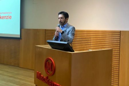 Imagem colorida mostra Ricardo Nunes, homem branco, de cabelo e barba pretos, vestindo terno cinza e camisa azul, no palco do auditório do Mackenzie. Ele está atrás de um púlpito de madeira com o logotipo de universidade, em um palco com paredes de madeira - Metrópoles