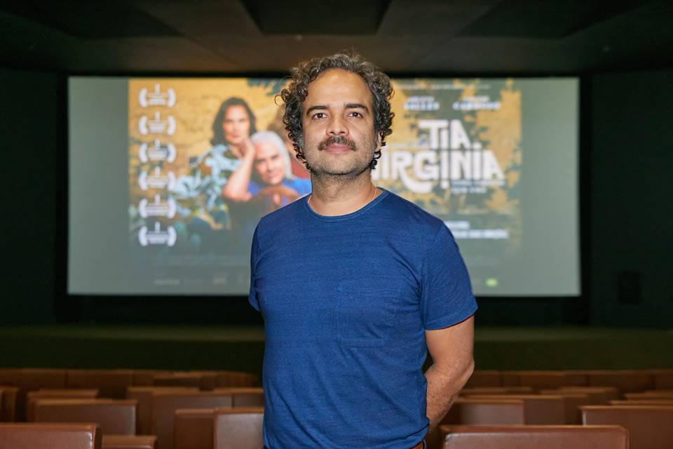 FOTO COLORIDA DE HOMEM POR VOLTA DOS 40 ANOS COM CAMISETA AZUL ESCURA DENTRO DE SALA DE CINEMA COM POSTER DO FILME TIA VIRGINIA AO FUNDO - METRÓPOLES