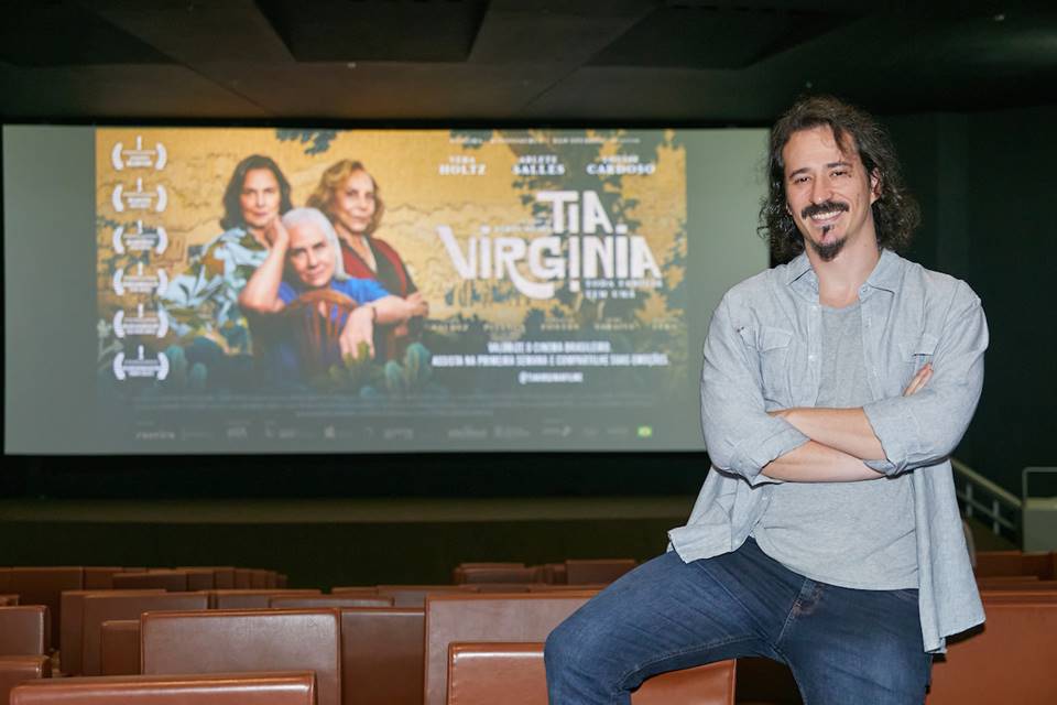 FOTO COLORIDA DE HOMEM JOVEM DE CAMISETA CINZA, CAMISA SOCIAL AZUL E JEANS DENTRO DE SALA DE CINEMA COM POSTER DO FILME TIA VIRGINIA AO FUNDO - METRÓPOLES