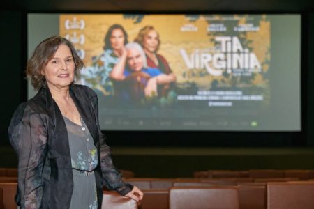 FOTO COLORIDA DA ATRIZ LOUISE CARDOSO DE VESTIDO ESCURO E CASACO PRETO DENTRO DE SALA DE CINEMA COM POSTER DO FILME TIA VIRGINIA AO FUNDO - METRÓPOLES