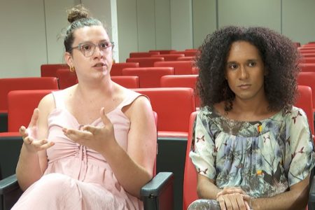 Imagem colorida mostra duas mulheres trans, uma delas com a pele branca, cabelo amarrado, usando óculos de grau e vestido rosa. A outra tem a pele parda, tem o cabelo escuro e solto, usando um vestido estampado em tons de cinza - Metrópoles