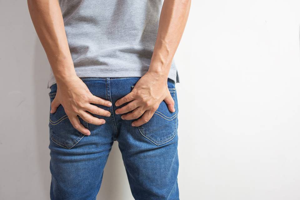 Foto colorida de um homem de costas da cintura para baixo de calça jeans e camiseta cinza, com as duas mãos nas nádegas em um fundo branco - Metrópoles