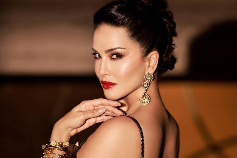 Foto colorida da atriz pornô Sunny Leone de costas olhando para a câmera, de batom vermelho e cabelo preso - Metrópoles
