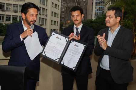 Imagem colorida mostra Ricardo Nunes, ao microfone, segurando uma pasta com um decreto assinado, ao lado de um empresário e do secretário Fabrício Cobra Arbex. Todos são homens, brancos, vestindo ternos escuros. eles estão em um terraço de um prédio do centro - Metrópoles