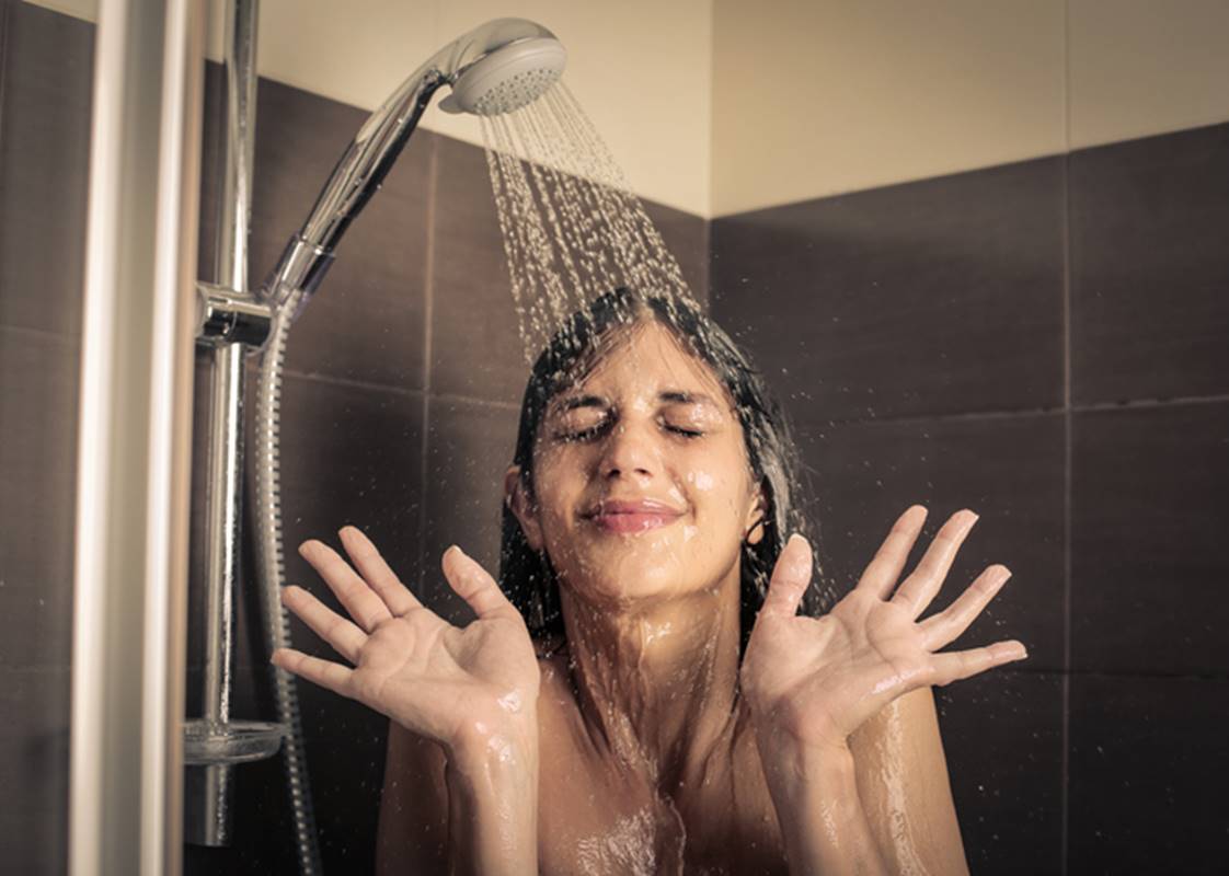 Imagem colorida de mulher mostrando as palmas das mãos, assustada, debaixo de um chuveiro, tomando banho frio - Metrópoles
