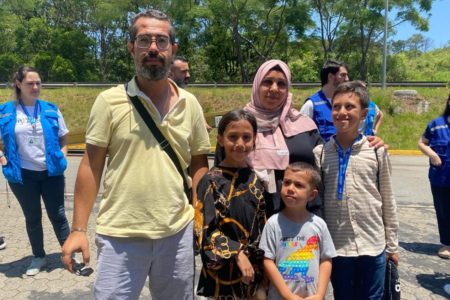 Imagem colorida mostra a família Bader, pai, mãe e três filhos, em pé, no sol, em um estacionamento com árvores ao fundo - Metrópoles