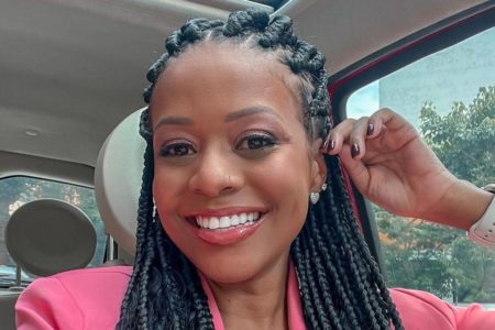 Foto colorida de uma mulher negra, com cabelo preto trançado, sorrindo, dentro de um carro, usando terno rosa - Metrópoles