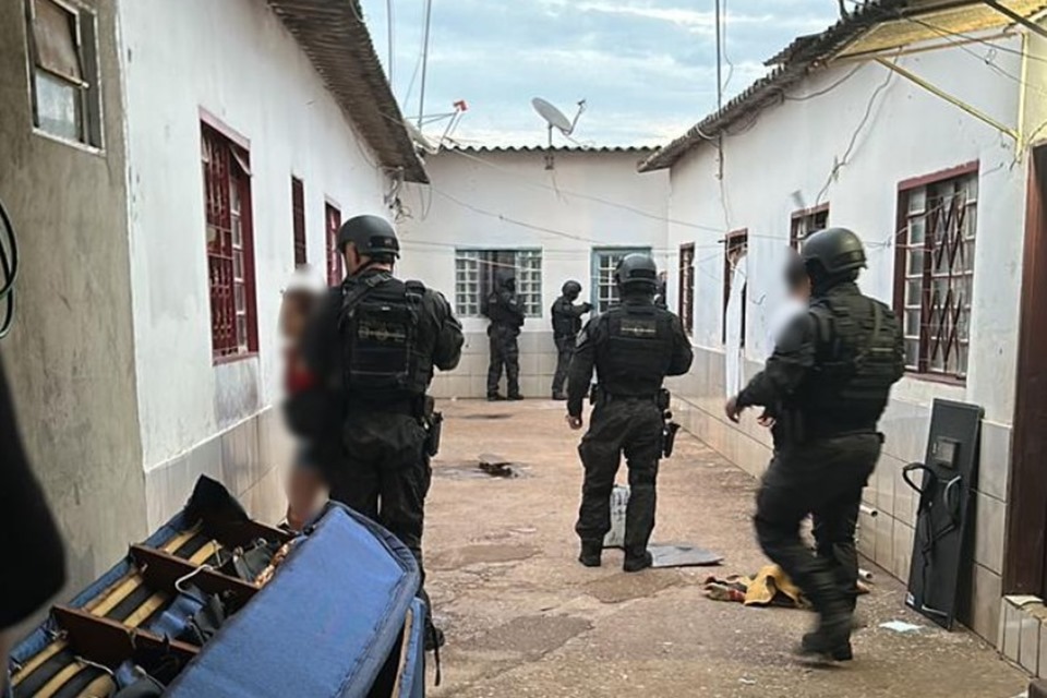 Foto colorida de policiais vestidos com uniformes pretos, em pequena rua cercada por casas brancas