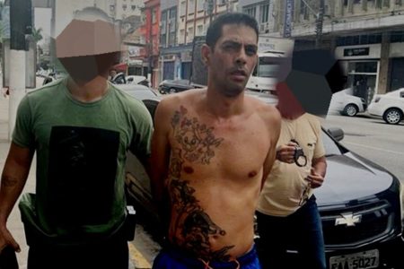 Em foto colorira homem sem camisa, tatuagens na lateral do corpo, é conduzido algemado, com mas mãos para trás, por dois policiais - Metrópoles