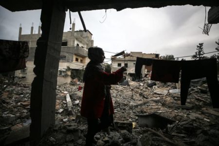 Imagem colorida mostra uma mulher recolhendo as roupas da própria casa, que foi destruída por bobmardeio - Metrópoles