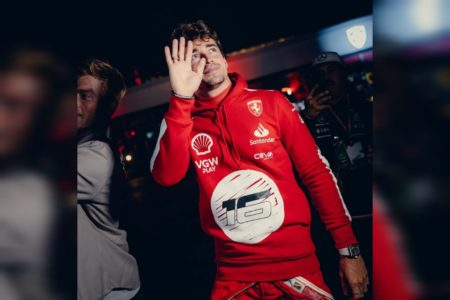 Foto mostra Charles Leclerc em primeiro plano usando um macacão automobilístico da Ferrari