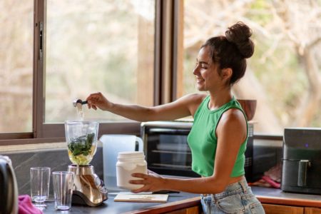 Jovem mulher preparando receita saudável em liquidificador com cozinha ao fundo - Metrópoles