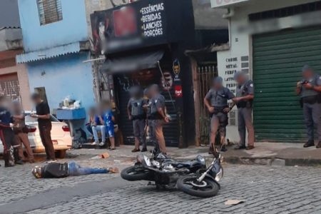 Foto colorida mostra jovem caído morto em via pública, ao lado de moto caída, observado por policiais militares e curiosos - Metrópoles