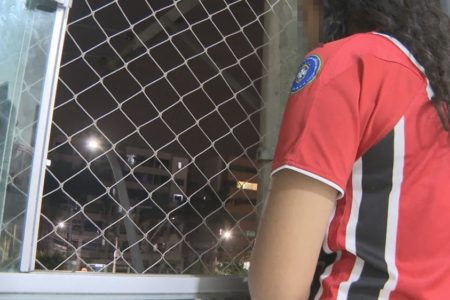 Foto colorida mostra pessoa de cabelos longos, cacheados, em frente a janela com tela de proteção, à noite; a pessoa está de costas para a câmera, vestida com camisa de time e tem o rosto borrado