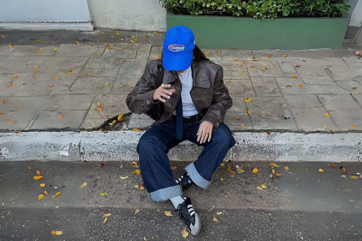 Sentada na calçada na rua, mulher usa boné azul, camiseta branca, calça jeans e tênis Adidas Samba - Metrópoles