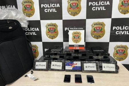 Foto colorida mostra tabletes de droga embalados em sacos pretos e celulares sobre mesa. Ao fundo, está um banner da Polícia Civil