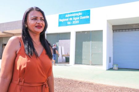 Foto colorida de mulher com cabelo liso e roupa marrom em frente à administração de Água Quente