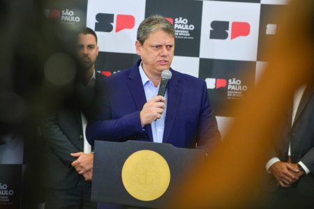 Imagem colorida mostra Tarcísio de Freitas, homem branco, grisalho, de terno azul escuro e camisa azul clara, falado ao microfone em um púlpito no palácio dos bandeirantes, entre câmeras - Metrópoles