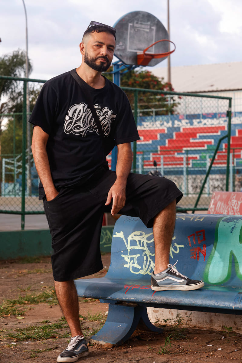 Homem com camiseta preta com grafiti colorido em branco, bermuda preta básica e calçados em azul com detalhes pretos e brancos.