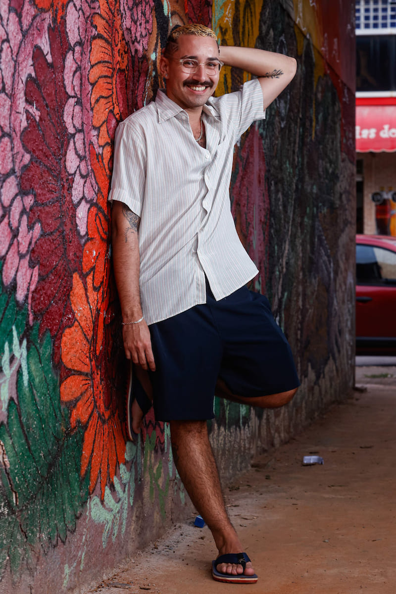 Homem vestindo uma camisa de botão listrada em tons claros, bermuda azul escuro e chinelos pretos com detalhes vermelhos.