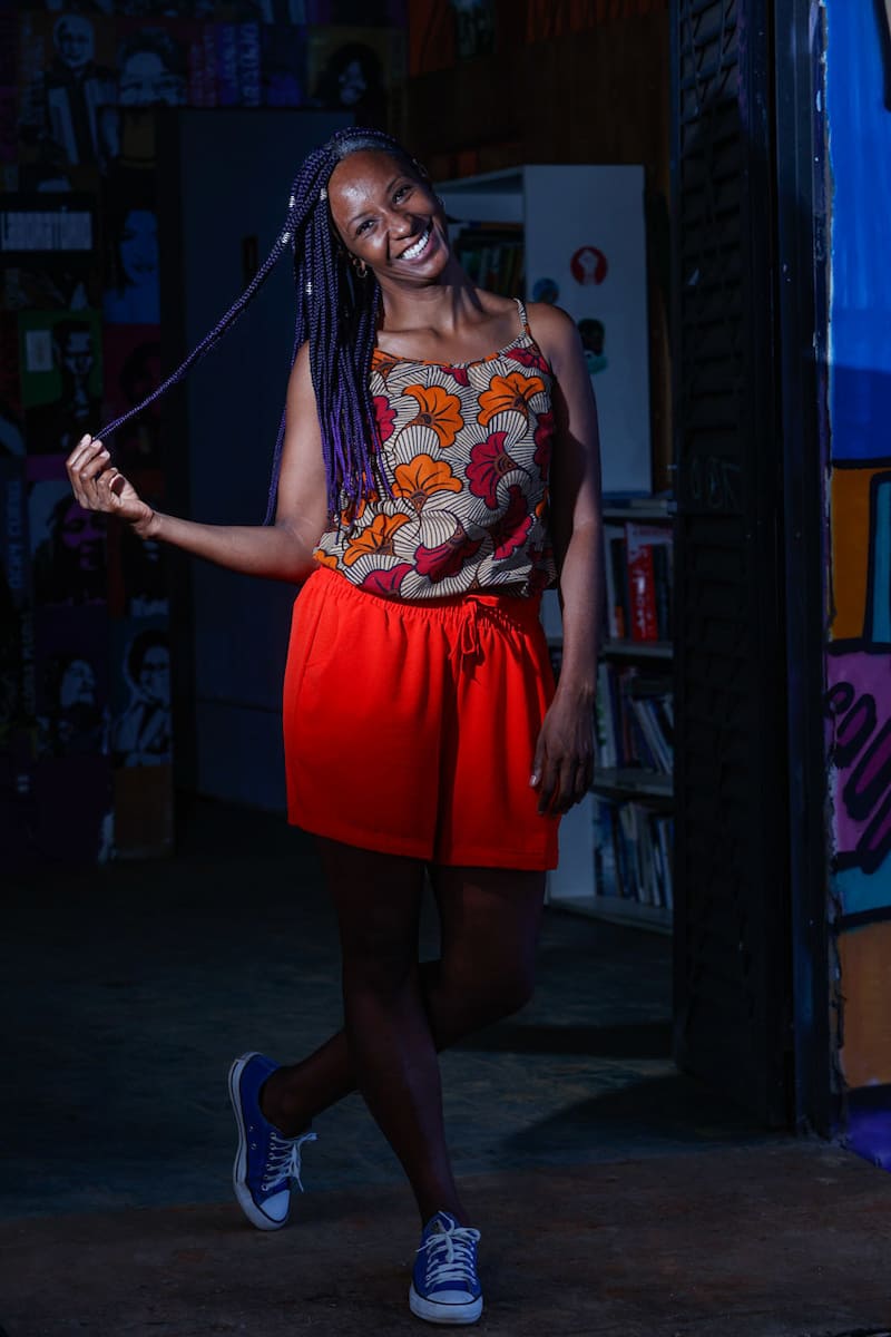 Mulher negra vestindo uma camiseta regata com um Kenté em vermelho e laranja junto a uma bermuda laranja e um calçado azul. 