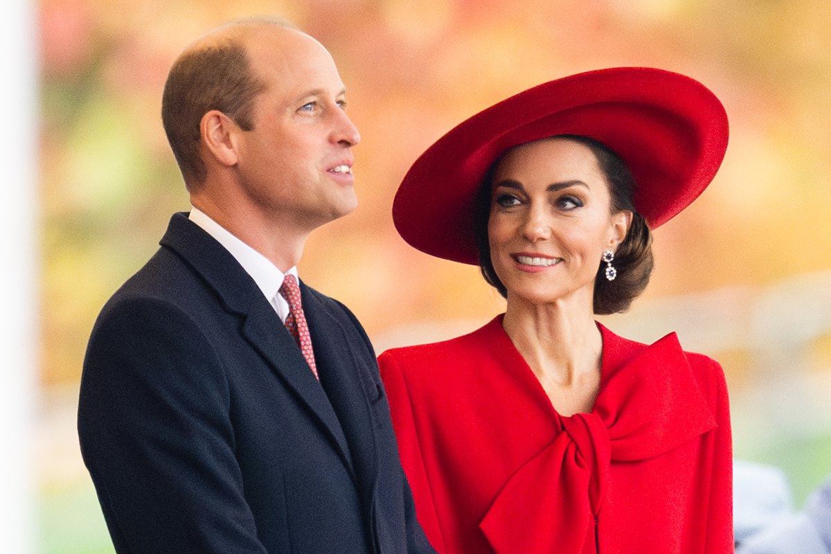 Foto colorida de homem calvo, olhando para cima, vestido com um blazer preto. Ele está ao lado de uma mulher de cabelo castanho preso. Ela usa um chapéu e uma roupa com laço grande na cor vermelha - Metrópoles