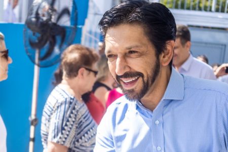 Imagem colorida mostra Ricardo Nunes sorrindo, em close do peito para cima. Ele é um homem branco, de meia idade, cabelo e barba pretos, e veste camisa azul. Ao fundo, há pessoas conversando - Metrópoles