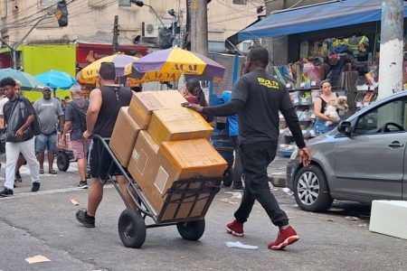 Imagem mostra homem puxando carrinho de mão, seguido por outro, em meio a uma rua com outras pessoas - Metrópoles