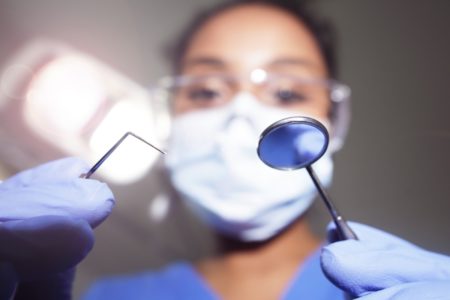 Imagem colorida mostra uma espátula e um espelho de dentistas, no primeiro plano, com uma dentista, mascarada, desfocada, ao fundo - Metrópoles