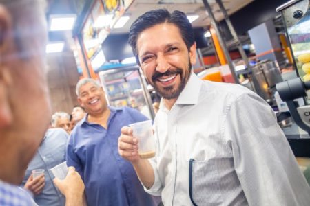 Imagem colorida mostra Ricardo Nunes, homem branco, de cabelo e barba pretos, vestindo uma camisa branca, segurando um copo de plástico de café com leite, em um bar popular repleto de pessoas sorrindo - Metrópoles