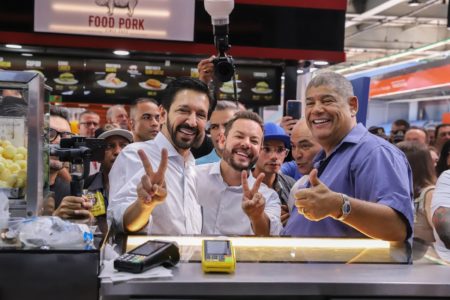 Imagem colorida mostra Ricardo Nunes fazendo um V com os dedos da mão direita, ao lado do presidente da Câmara, Milton Leite, fazendo sinal de positivo. Ambos sorriem. Nunes está de camiseta branca. Leite, de camiseta polo azul. A imagem é da cintura para cima. Eles estão em um balcão de uma lanchonete um terminal de ônibus lotado da zona sul; prefeito anunciou a chamada Tarifa Zero aos domingos na capital paulista - Metrópoles