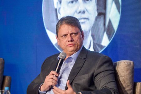 Fotografia colorida mostra Tarcísio de Freitas, homem grisalho, olhando para o lado enquanto segura um microfone com a mão direita em frente a um fundo azul com a sua fotografia - Metrópoles