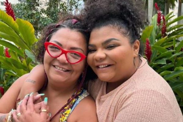 Foto colorida de Babi Cruz com a filha Flora Cruz, ao fundo, folhas verdes - Metrópoles