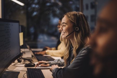 Imagem colorida de mulher loira, sorrindo, durante um atendimento em call center. Ela usa um microfone e olha para a tela do computador - Metrópoles